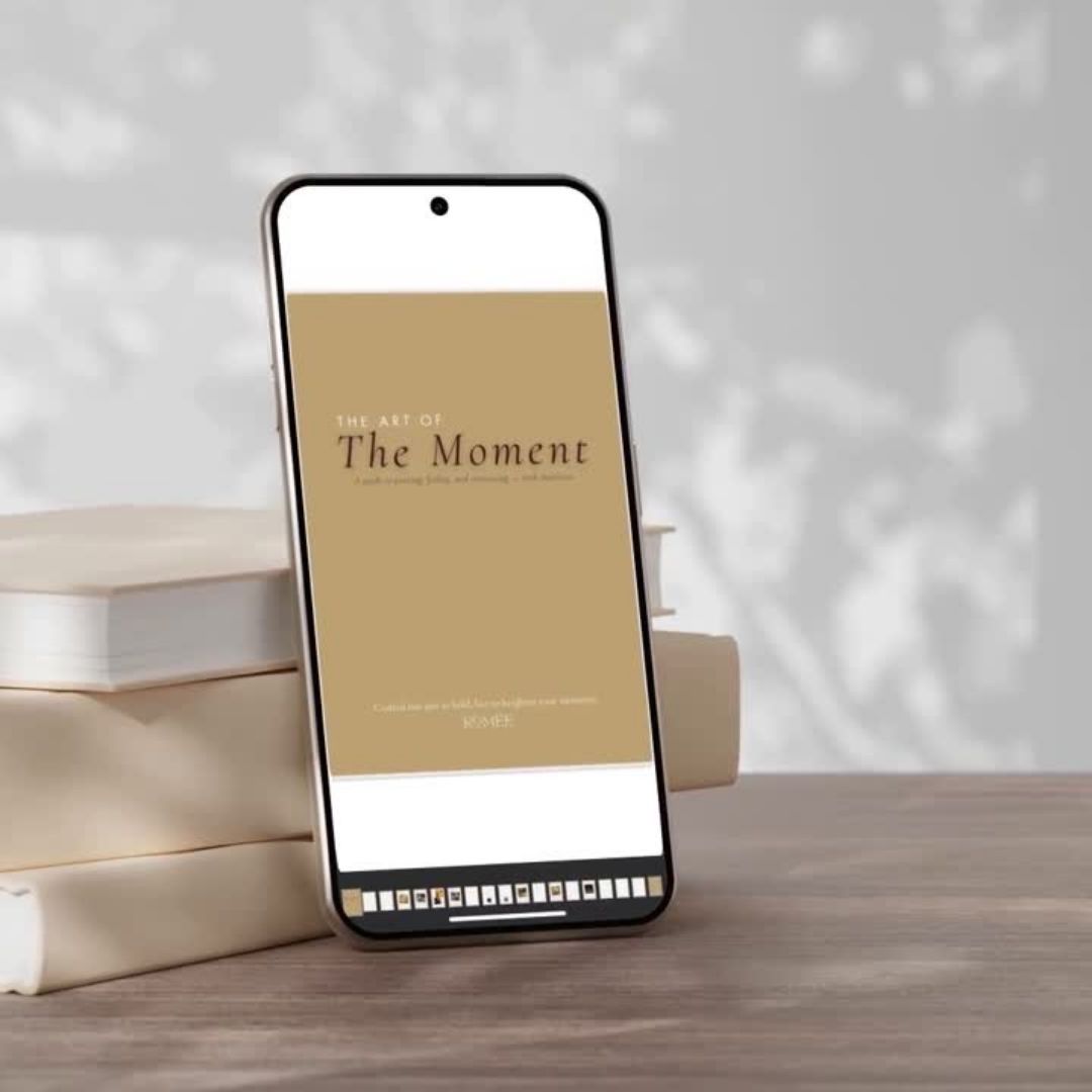 Phone displaying 'The Art of the Moment' e-book cover on a wooden surface with books in the background